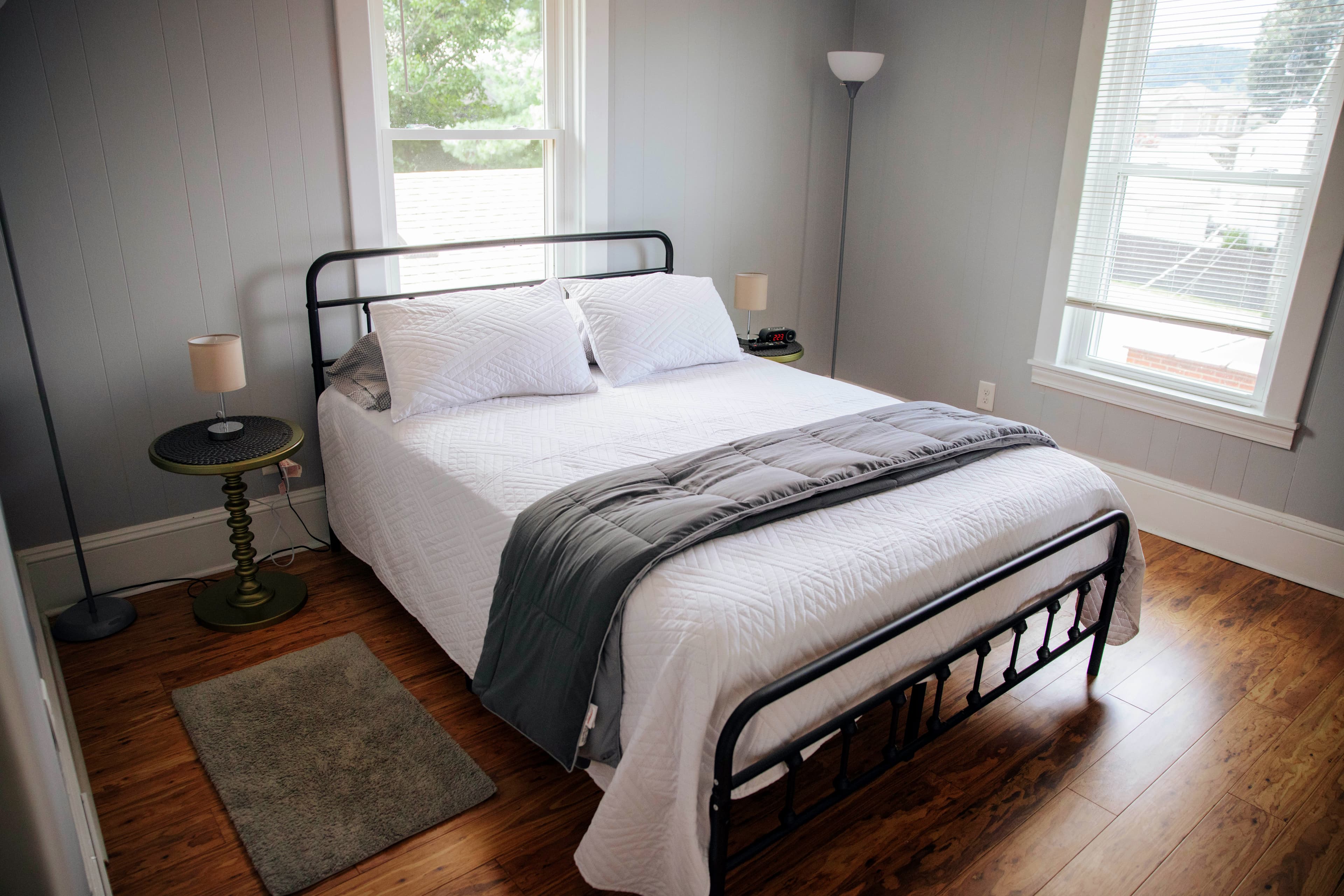 A cozy bedroom featuring a metal frame bed, gray bedding, and natural light from the windows.