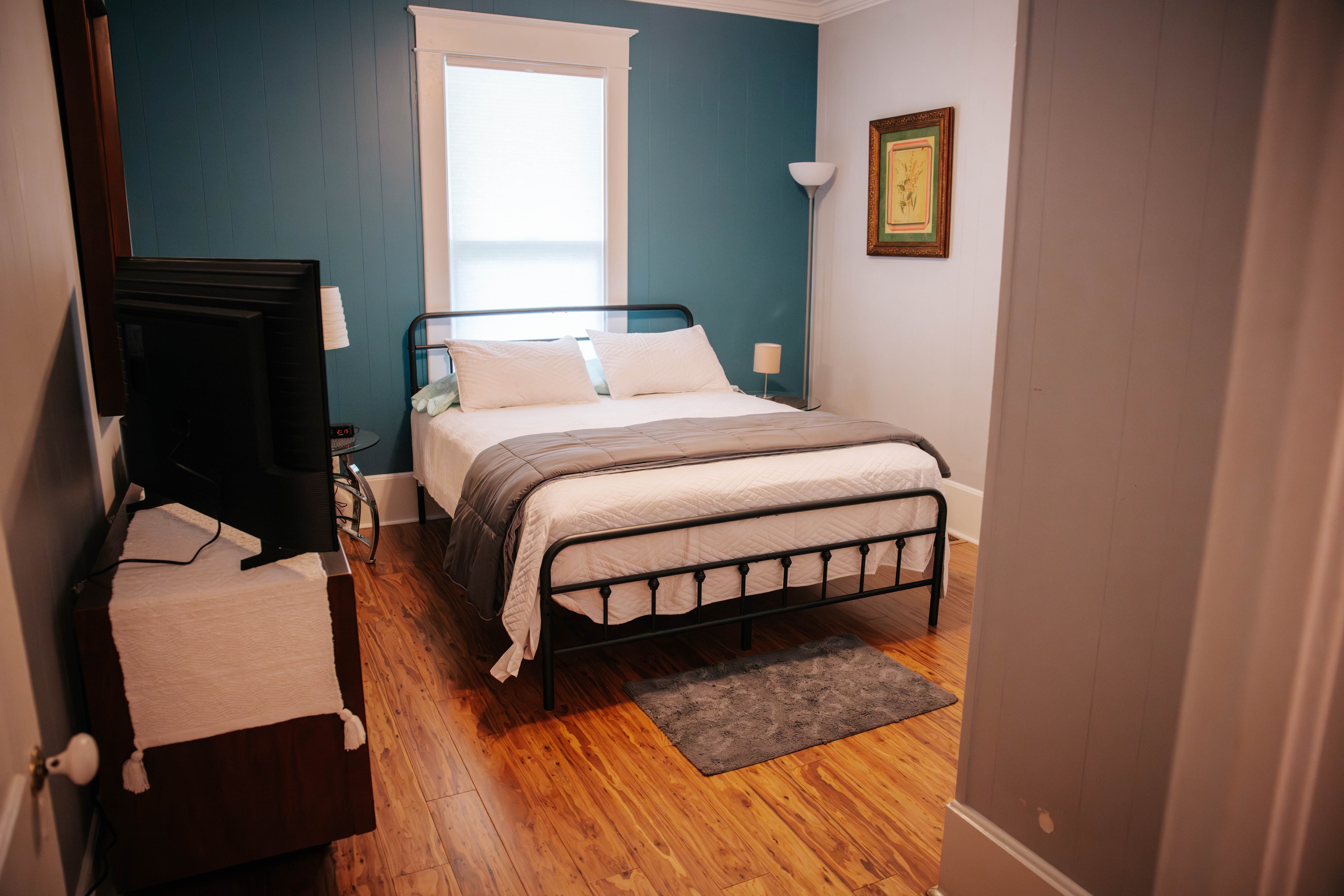 A cozy bedroom featuring a metal bed frame, wooden flooring, a TV, and teal walls.