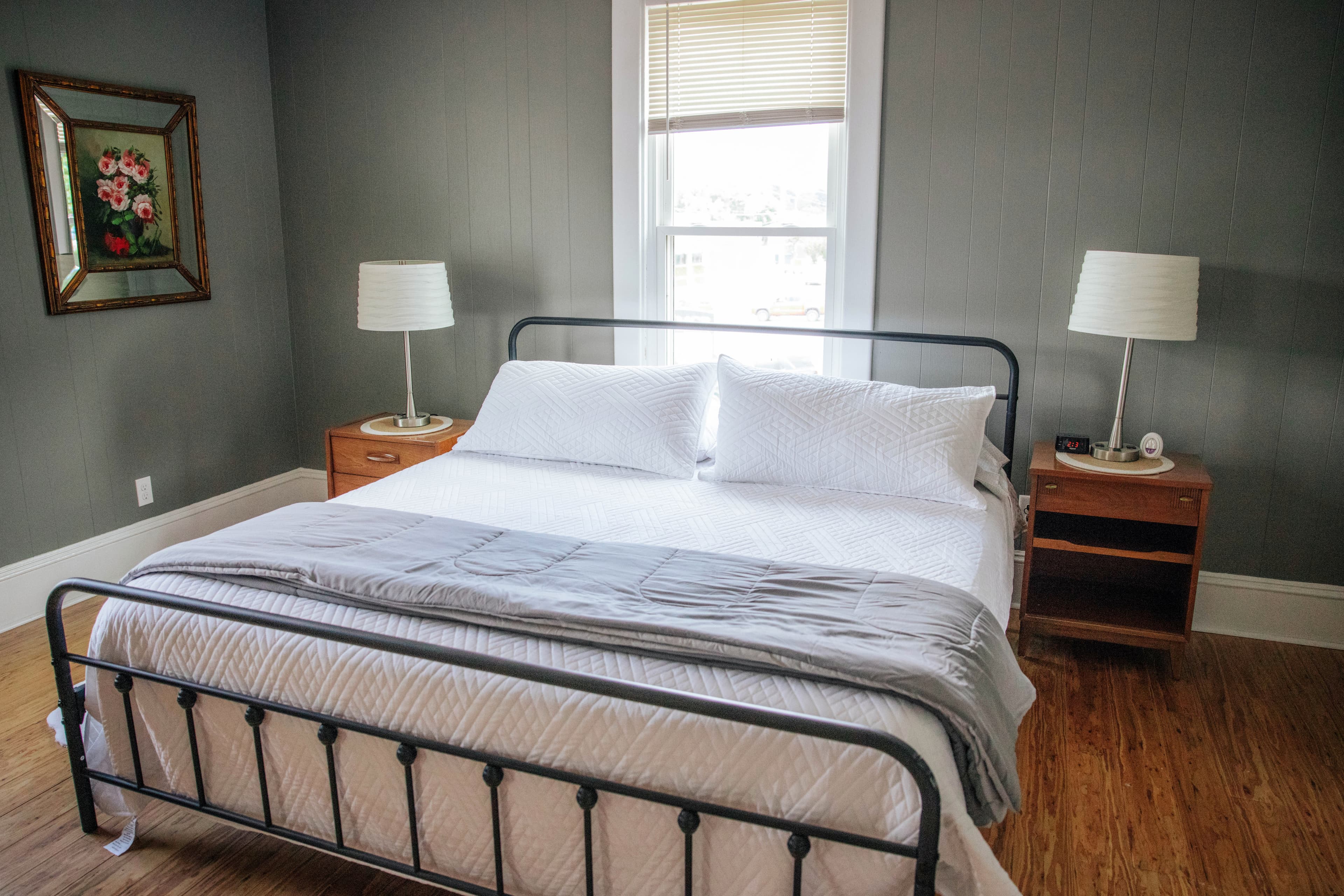 A neatly made bed with white linens is centered in a cozy, softly lit bedroom featuring gray walls and wooden floors.