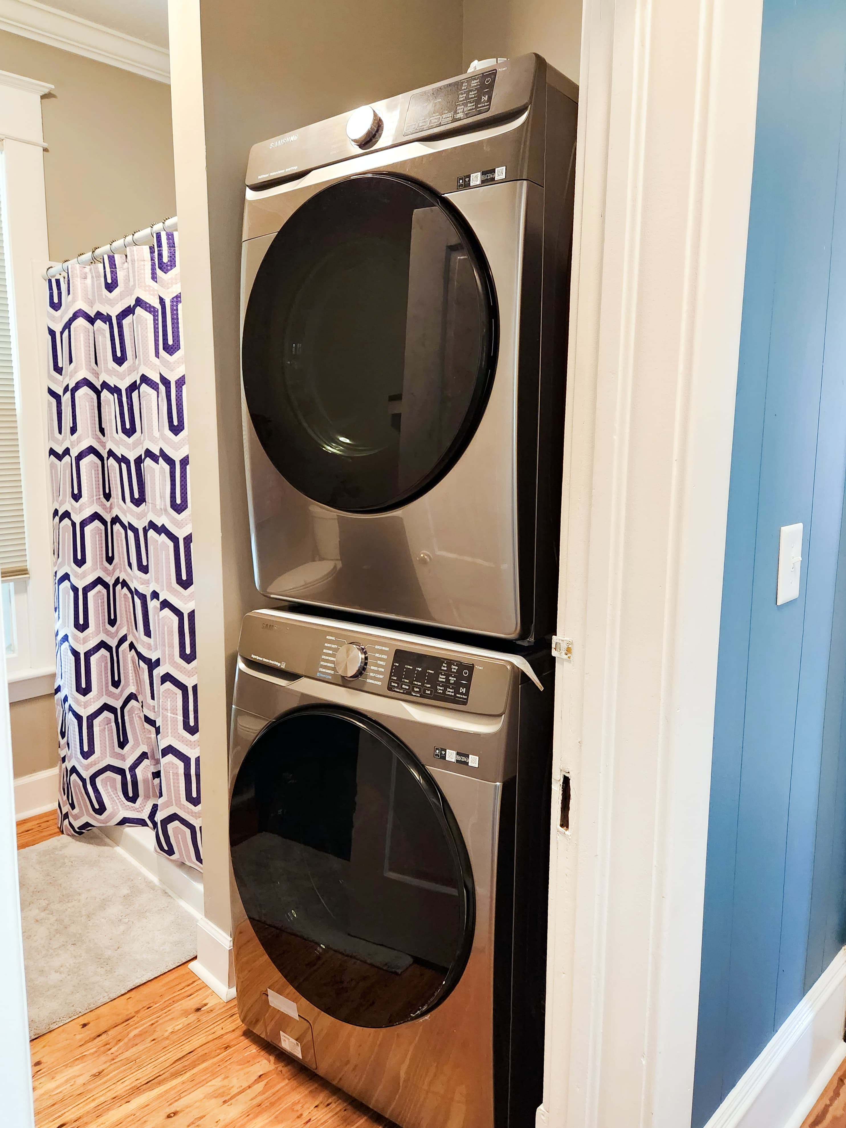 A stacked washer and dryer in a narrow laundry space next to a patterned shower curtain.