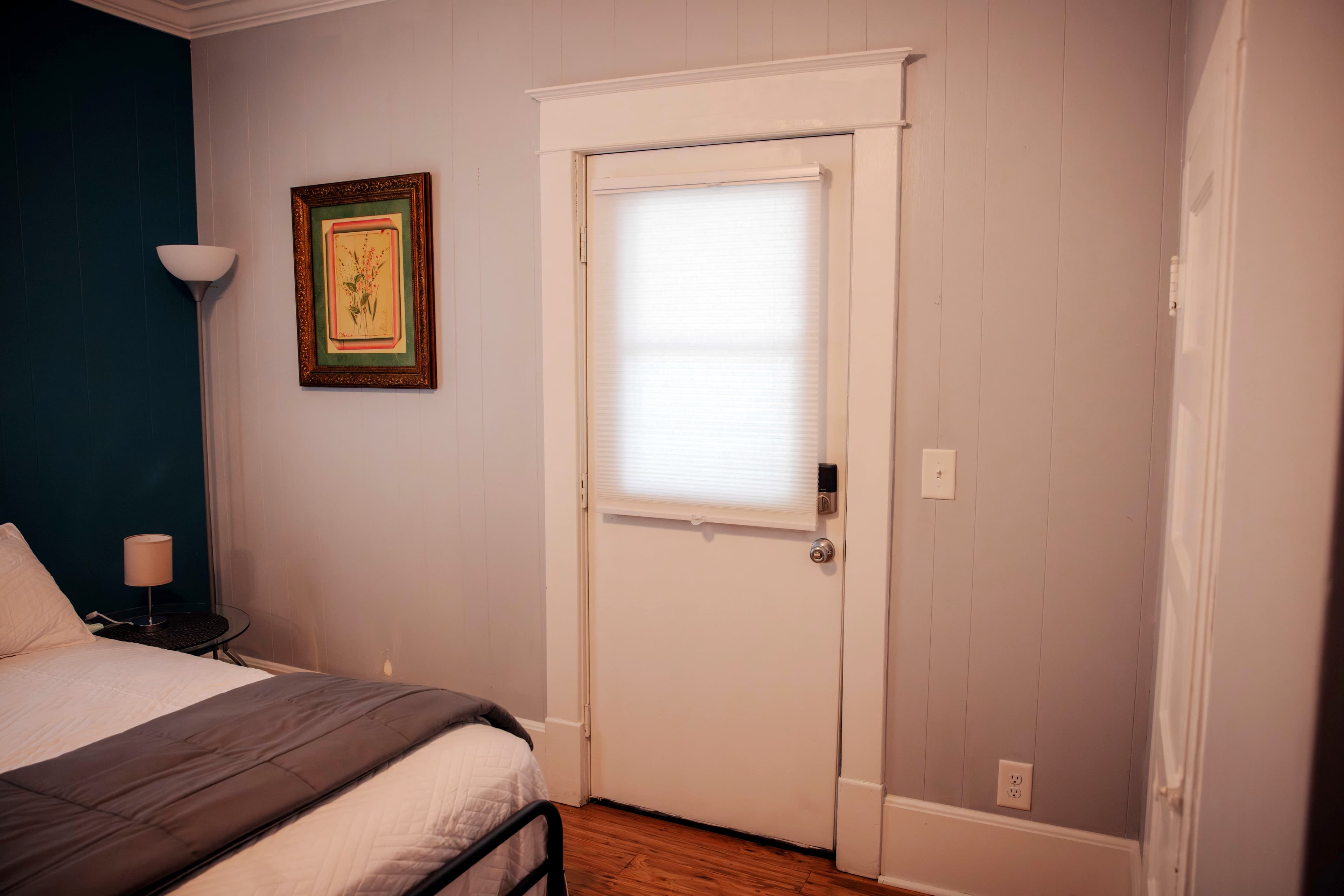 A cozy bedroom featuring a white door with a window, framed artwork on the wall, and a bedside lamp.