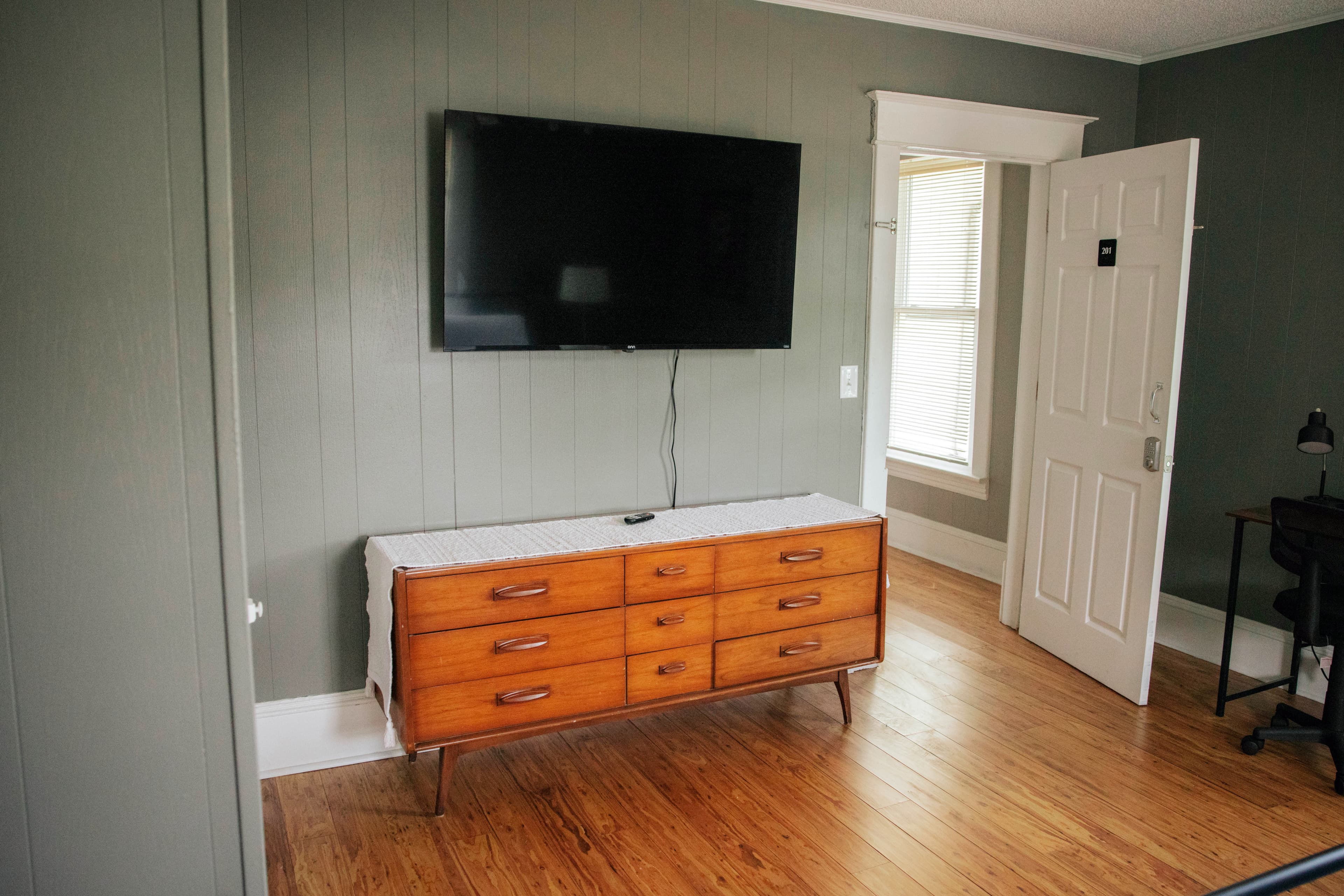 A simple room featuring a wooden dresser, a mounted TV on the wall, and a door leading to an outside area.