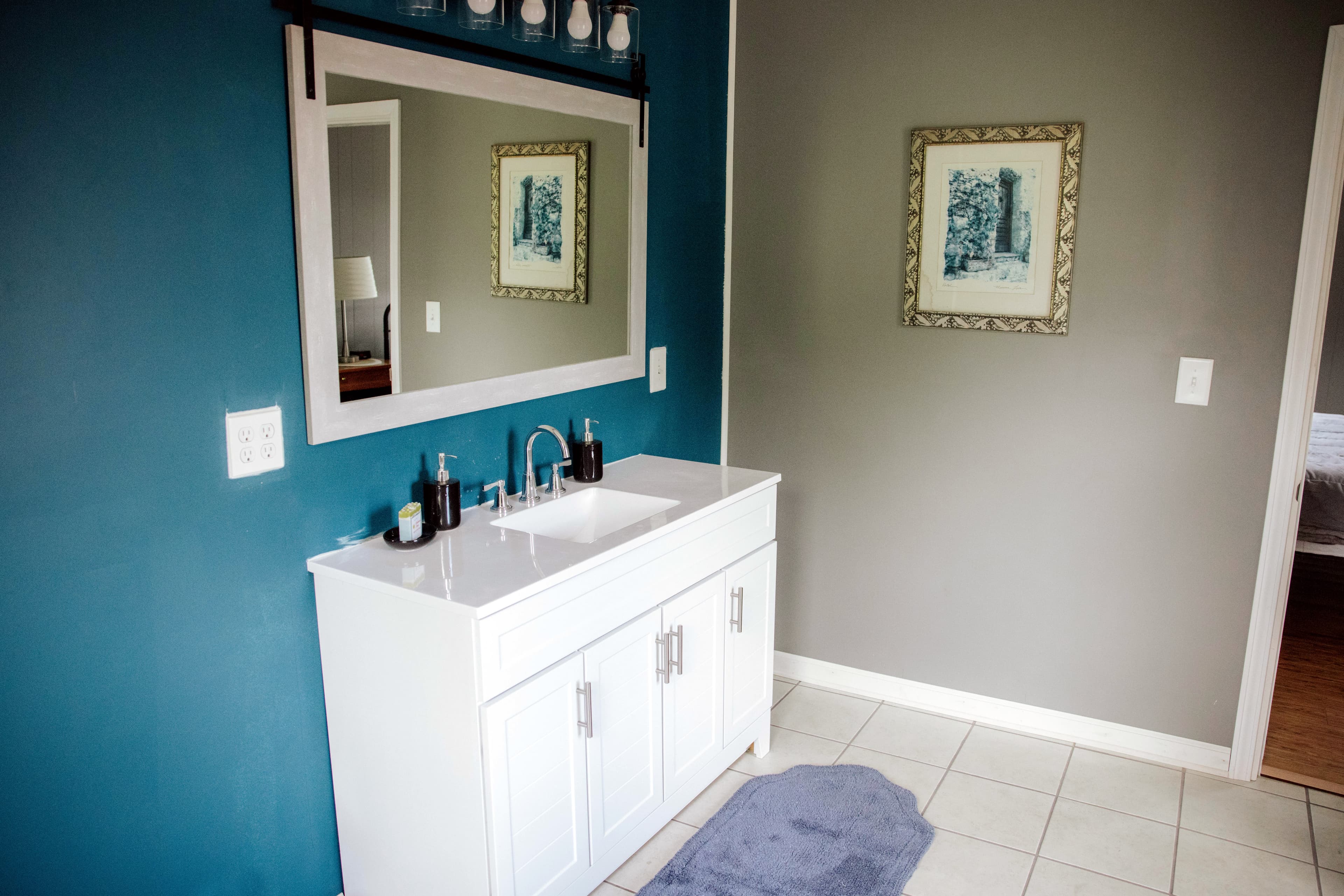 A modern bathroom with a white vanity, mirror, and teal walls, featuring decorative artwork and a plush rug.