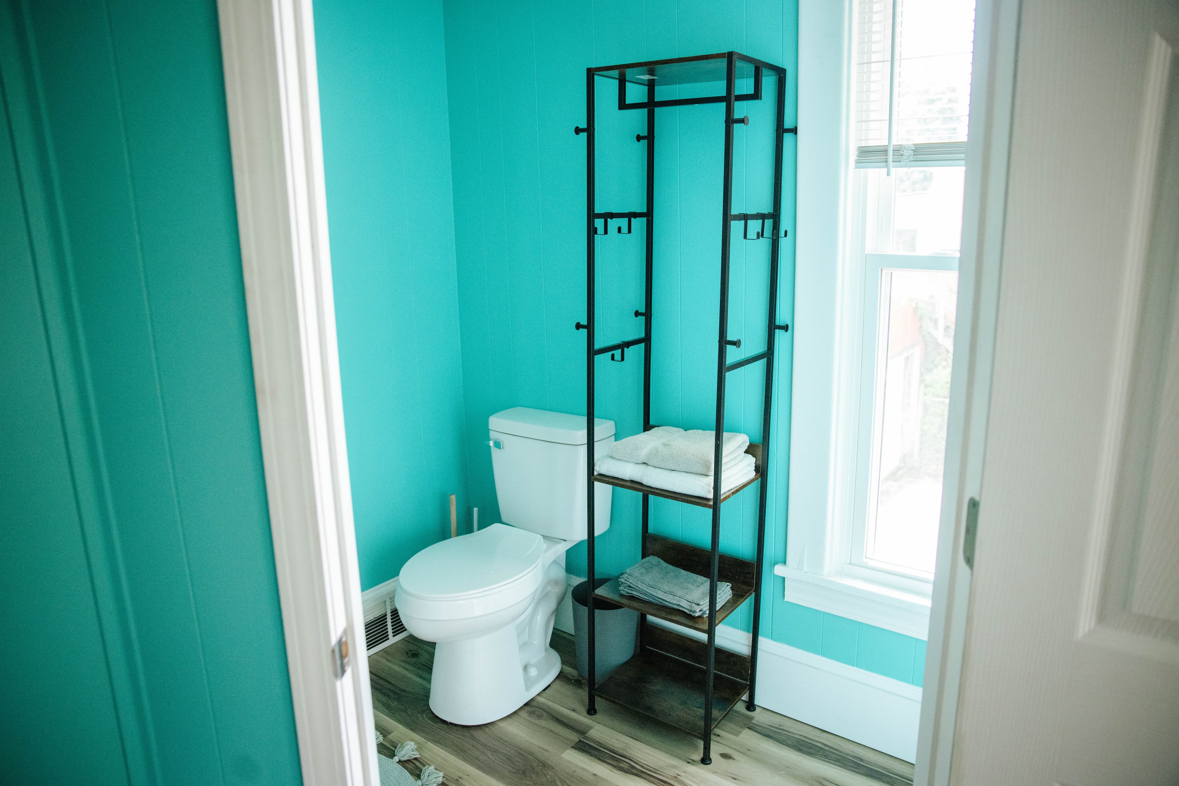 A bright turquoise bathroom featuring a toilet and a metal shelving unit with towels.