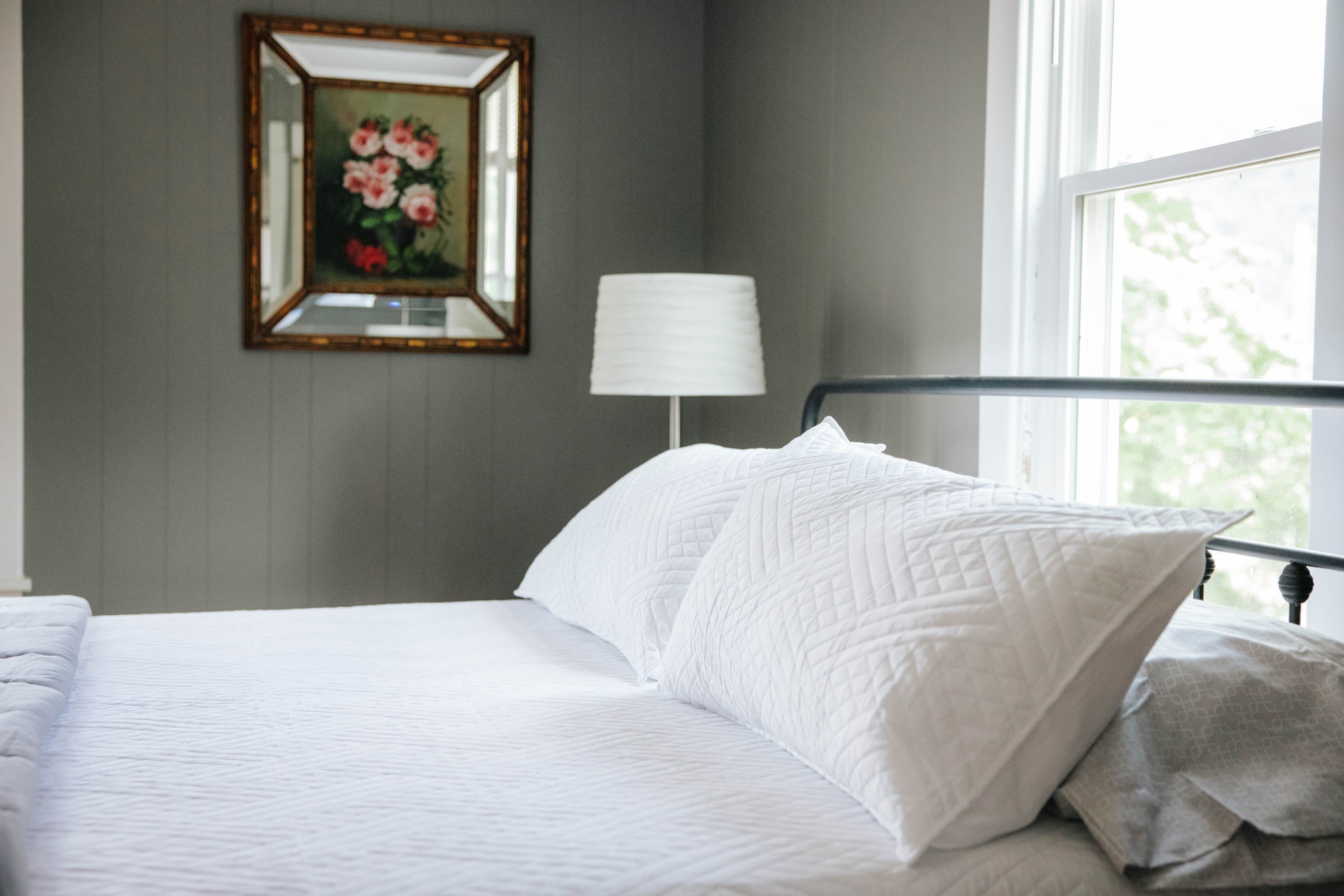 A cozy bedroom featuring a bed with white pillows, a lamp, and a floral painting on the wall.