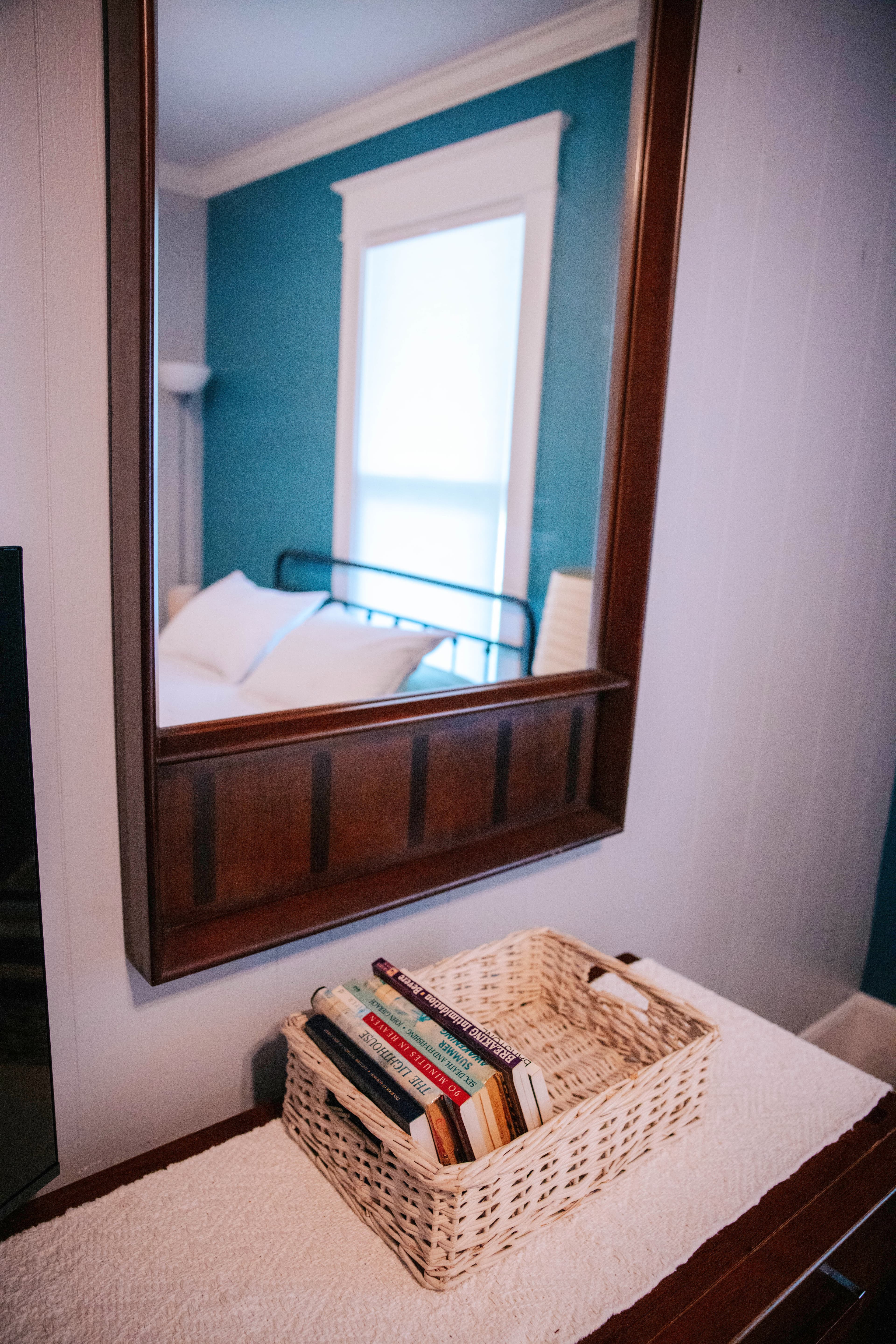 A wooden mirror reflects a bright blue wall and a tidy bed, alongside a woven basket filled with books on a white surface.