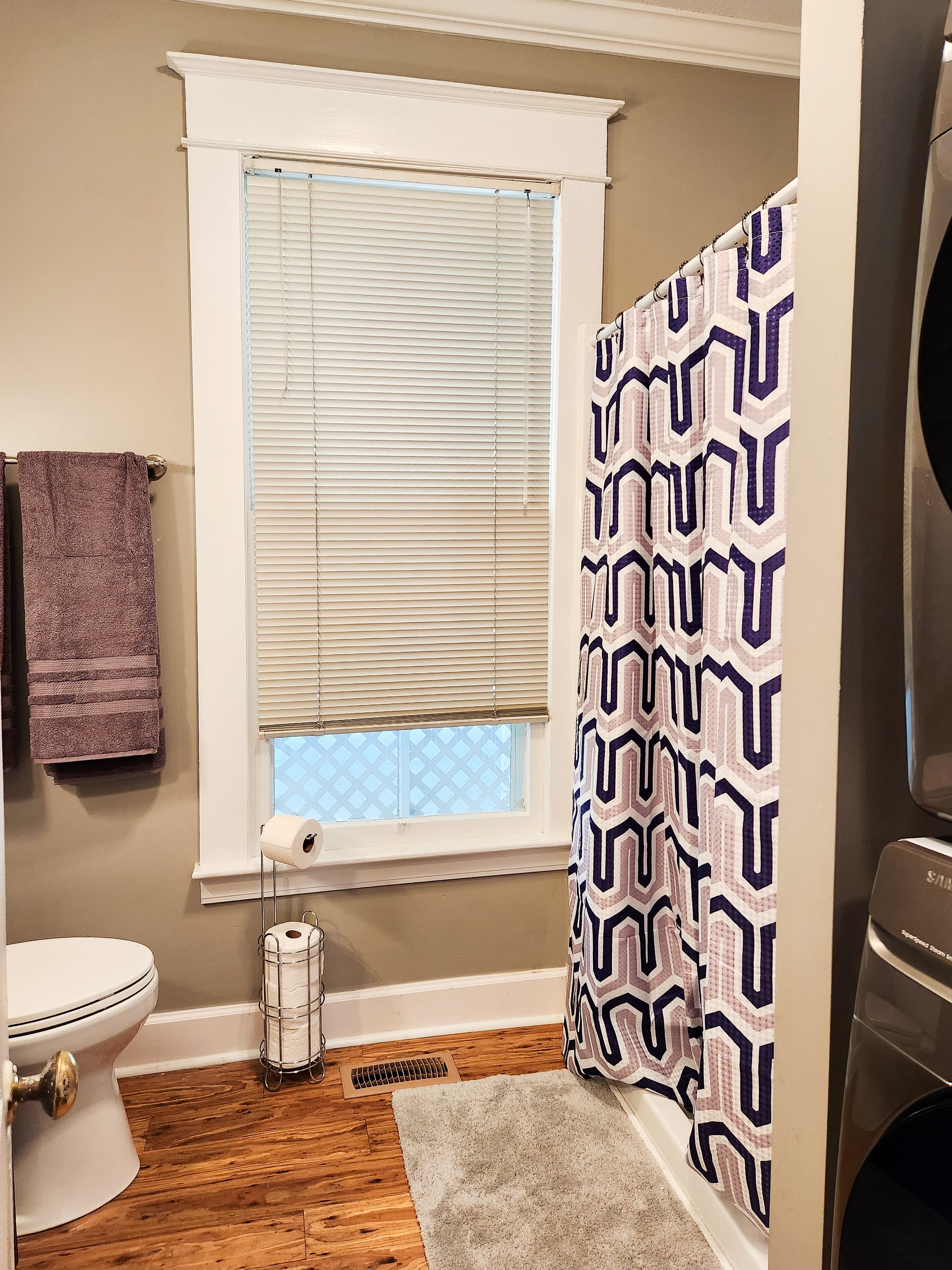 A small bathroom featuring a shower with a patterned curtain, a toilet, and a window with blinds.